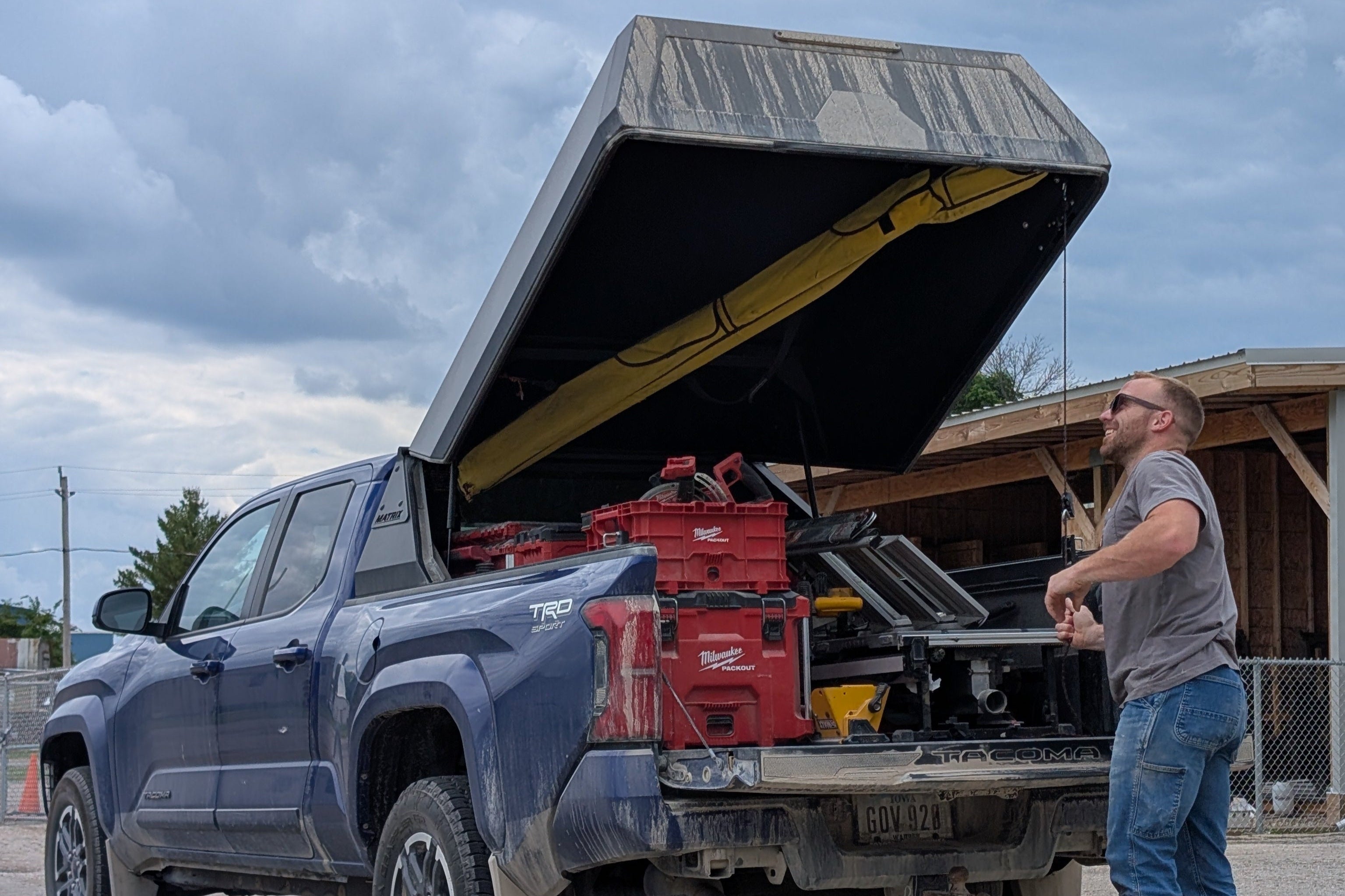 Man standing next to a pickup truck with an open bed and tools inside, under a cloudy sky.